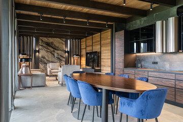 large modern kitchen and living room. Large wooden dining table, blue chairs, wooden panels on walls. Wooden kitchen furniture, black countertop, steel extractor hood. Grey armchairs, floor lamp, TV.
