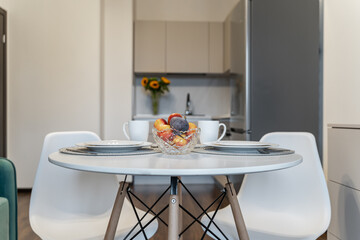 design of simple modern kitchen. bright kitchen. round white table and chairs, fruit dish on table, plates, cups for coffee or tea, yellow sunflowers in vase in background. Modern flat. inside home