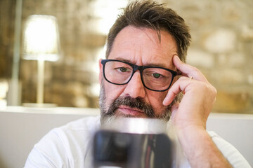 Man sitting at home, looking at his phone with a serious expression while reading a phishing scam notification. He struggles to comprehend the message, feeling confusion as he browses the web 
