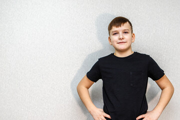 A boy in a black T-shirt stands against a grey wall with his hands on his hips.