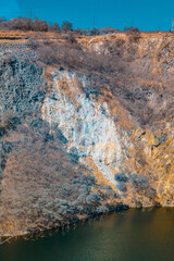 A serene, almost surreal landscape photograph depicts a rocky hillside, partially bare of vegetation, sloping down to a dark, still body of water under a clear blue sky