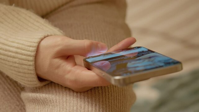 Woman comfortably wearing sweater, swiping smartphone screen while scrolling social media news feed in cozy, light filled home environment