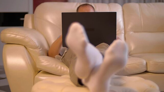 A man sits comfortably on a white leather sofa with his feet on an ottoman in a large room and works absorbedly on a black laptop. Shop in motion