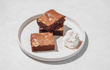 Chocolate brownie with nuts cut into pieces on a plate with whipped cream on a light background