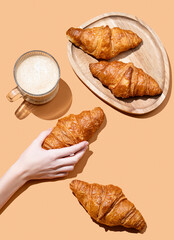 Hand holding fresh croissant on orange background with cappuccino cup and fresh pastry