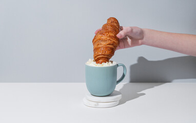 Hand dipping fresh croissant into cup of coffee with whipped cream on gray background with shadow.