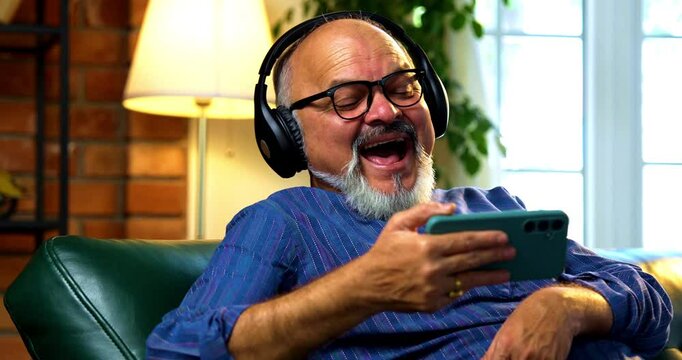 Indian senior retired man with beard wearing kurta laughing out loud while watching funny video on smartphone with wireless headphones, enjoying entertainment