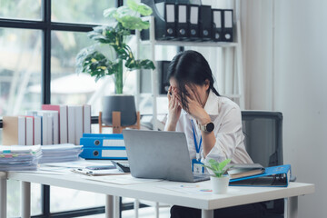 Overwhelmed Office Worker: A young businesswoman sits at her cluttered desk, hands covering her...