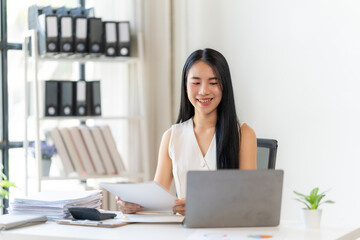 Confident Businesswoman at Work: A young, smiling businesswoman sits at her desk, reviewing documents and working on her laptop. She exudes confidence and professionalism in her modern office setting.