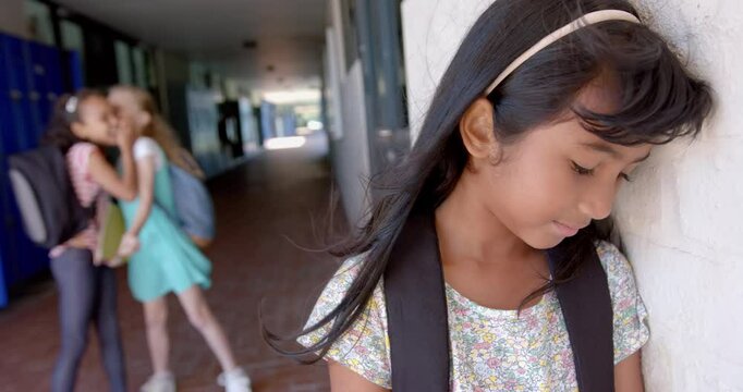 In school, girl with backpack feeling sad while other girls whispering nearby