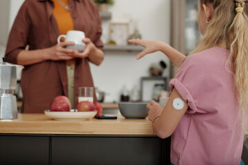 Closeup of young girl with diabetes talking to mother in kitchen setting with focus on CGM device...