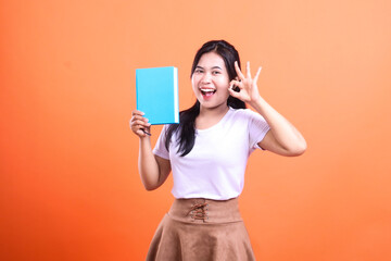 Naklejka premium A young woman with dark hair holds a blue book in one hand and gives a hand okay sign in the other hand