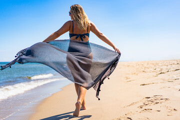 Beautiful middle aged woman holding scarf fluttering in wind walking on seashore on sandy beach....