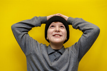 boy ten years old in black knitted hat looks up with great surprise and joy on yellow background....