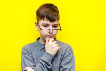 A ten year old boy in rectangular glasses stands thoughtfully with his hand to his chin on a yellow background.