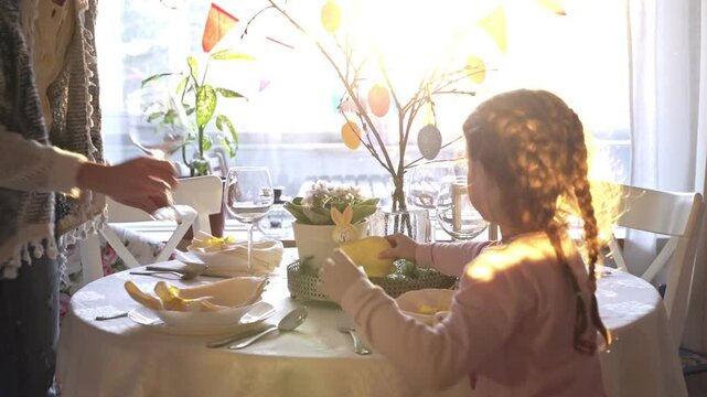 Little girl and her mom serve festive Easter table with a yellow bunny decoration. Sunlight fills the cozy room, elegant tableware, pastel colors, and holiday decor. A warm family moment full of joy
