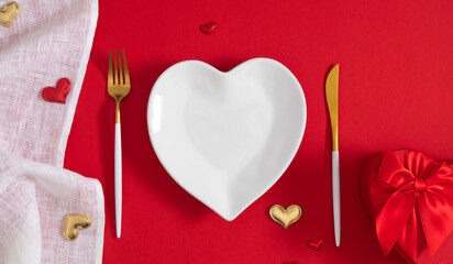 Beautiful festive background for Valentine's Day. White heart-shaped plate, knife and fork, heart confetti, white linen tablecloth and gift on red background. Valentine's Day concept. Flat lay