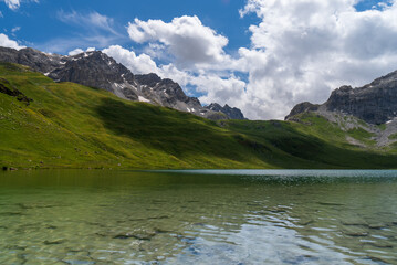 Obraz premium Serene beauty of Savoie in the French Alps with this stunning photograph. Featuring lush green meadows, a winding stream, and towering snow-capped peaks against a clear blue sky