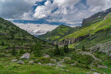 Naklejka premium Breathtaking mountain landscape in Savoie, French Alps. Snow-capped peaks, lush meadows, rocky cliffs, and a cascading waterfall under a dramatic sky.