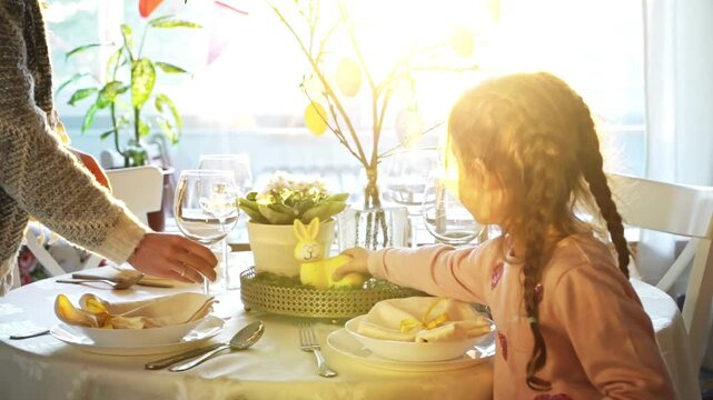Little girl and her mom serve festive Easter table with a yellow bunny decoration. Sunlight fills the cozy room, elegant tableware, pastel colors, and holiday decor. A warm family moment full of joy