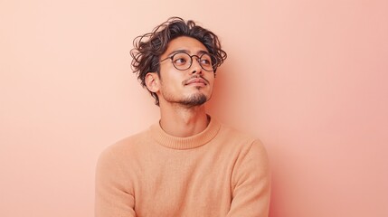 Young man in glasses looking up wearing peach sweater against pink wall.