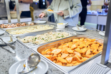 Close-up of a metal container with baked hearty food. A woman puts food in a cup on a buffet table...