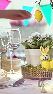 A young woman serves a festive Easter table with a yellow bunny decoration. Sunlight fills the cozy room, highlighting elegant tableware, pastel colors, and holiday decor. A warm family moment full of
