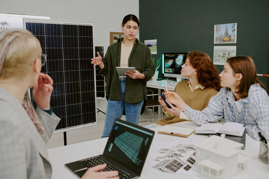 Woman presenting renewable energy project to diverse team using a tablet and solar panel display. Group of colleagues listening attentively and discussing technical details
