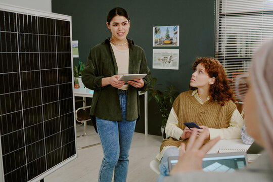 Young woman presenting sustainable energy ideas using tablet in modern office setting with colleagues listening attentively, surrounded by solar panel and green decor