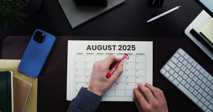 A man highlights dates on his calendar while sitting at his desk in the office. Close-up of his hands, top view