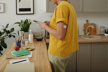 Side view cropped shot of woman with diabetes using smartphone standing in kitchen with focus on...