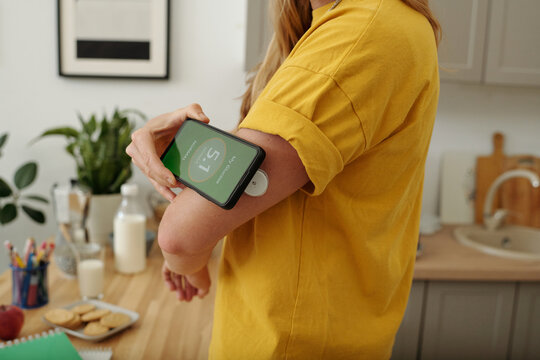 Closeup of unrecognizable woman with diabetes putting smartphone to CGM device on arm while monitoring glucose levels at home screen showing normal green readings copy space
