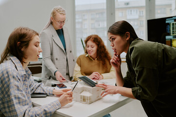 Group of diverse colleagues engaging in collaborative discussion around office table with various devices. Bright office space with large windows and city view in background