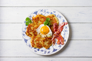 Hash browns with fried egg and bacon on white plate and white wooden background. View from above, top view