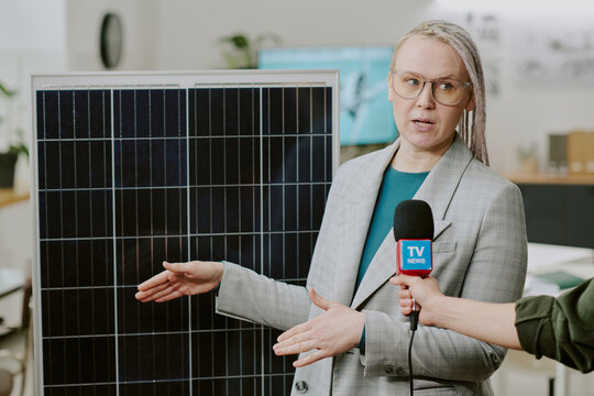 Woman in business attire explaining solar panel benefits during an interview in office environment She is holding microphone and standing near solar panel