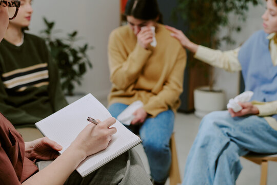 Group of individuals sitting in circle engaging in therapy. One individual writing in notebook while another comforts a crying participant using a tissue