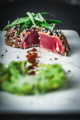 Close-up of a delicious tuna steak in a sesame crust. Medium-rare tuna slices with green puree and sun-dried tomatoes on a white plate on a dark background. Vertical photo.