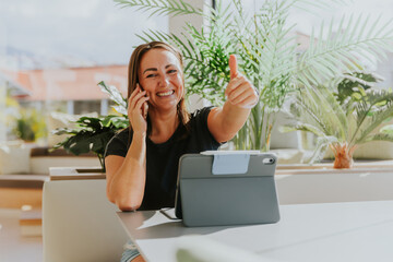 A woman is having a phone call and giving the thumbs up while working in the office, positive vibe