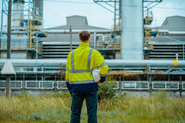 Worker observing industrial plant operations while standing in a grassy area during daylight hours