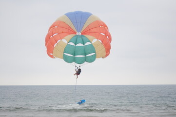 A brightly colored parasail is flying high above the ocean. The parasailer is harnessed in and enjoying the view. A small boat is visible in the water below.