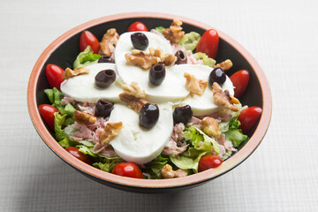 Bowl of fresh mozzarella salad with lettuce, tomatoes, olives, walnuts and ham over a white background