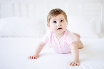 Fototapeta premium a six-month-old baby learns to crawl on a white cotton bed at home, a little baby girl in a pink bodysuit smiles, close-up