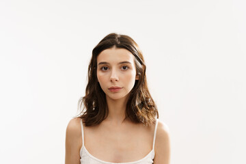 Fototapeta premium Young brunette woman in a white light top posing against a white background, expressing emotions and touching her face with her hands. Minimalistic and natural beauty concept