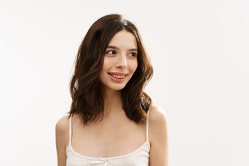 Fototapeta premium Young brunette woman in a white light top posing against a white background, expressing emotions and touching her face with her hands. Minimalistic and natural beauty concept
