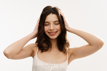 Fototapeta premium Young brunette woman in a white light top posing against a white background, expressing emotions and touching her face with her hands. Minimalistic and natural beauty concept