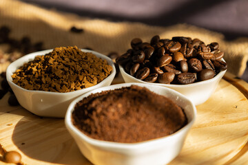 Focus of instant coffee and coffee beans in white porcelain bowls, on a wooden plate and scattered coffee beans on a dark background illuminated by the morning sun