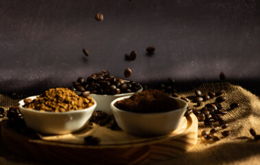 Coffee beans falling into small porcelain bowls with different types of coffee. beans, ground and instant, on a wooden plate on a dark background with sunlight coming through the window