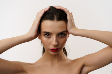 Fototapeta premium Young brunette woman with red lipstick posing against a white background, expressing emotions, and touching her face with her hands. Minimalistic beauty and fashion concept