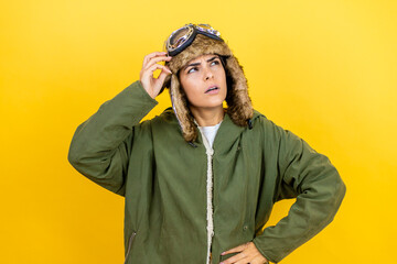Young beautiful aviator woman wearing vintage pilot helmet with glasses and jacket over yellow background confuse and wonder about question. Uncertain with doubt, thinking with hand on head
