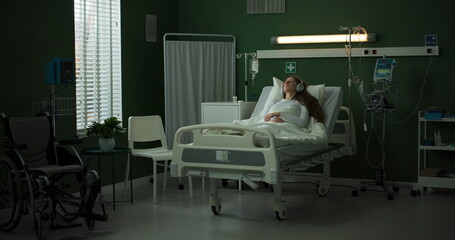 Resting in a hospital bed, a woman listens with headphones, eyes closed.